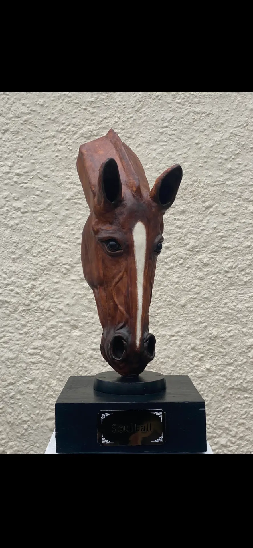 Hand-sculpted chestnut horse-head sculpture Soul Fall on a black plinth against a stone wall, with brass name plaque